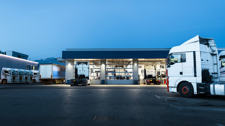 A modern truck service center at dusk with several white semi-trucks parked outside and others being serviced inside the brightly lit bays.