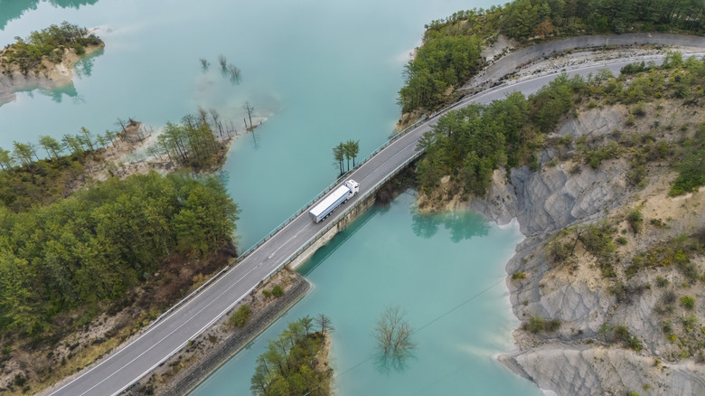 A semi-truck travels across a narrow bridge spanning a bright turquoise lake.