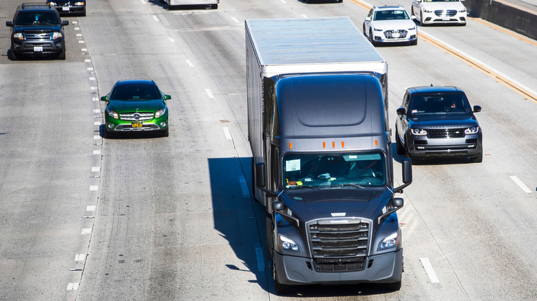 A black semi-truck drives in heavy freeway traffic surrounded by cars.