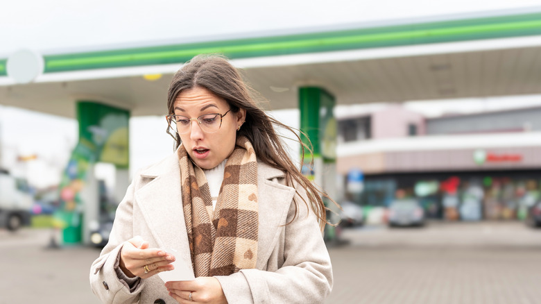 A young woman in a white coat outside a BP gas station looking stressed as she reads a receipt