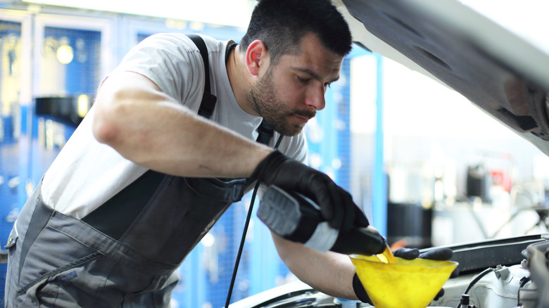 A mechanic wearing black overalls pours oil into a yellow funnel.