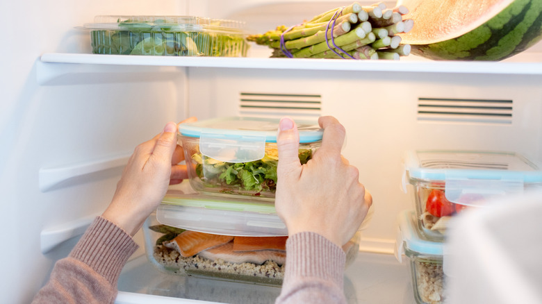 A person holding either side of a lidded container in a fridge filled with produce and some leftovers.