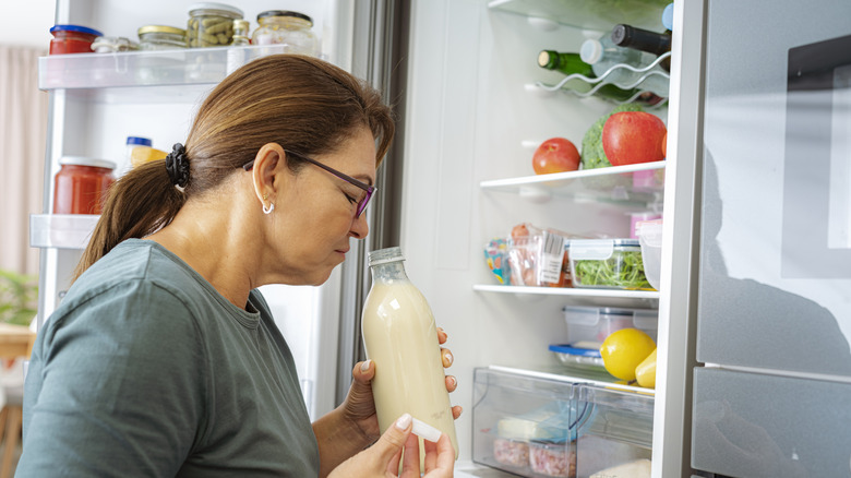 A woman smelling an open bottle of milk in front of a refrigerator.