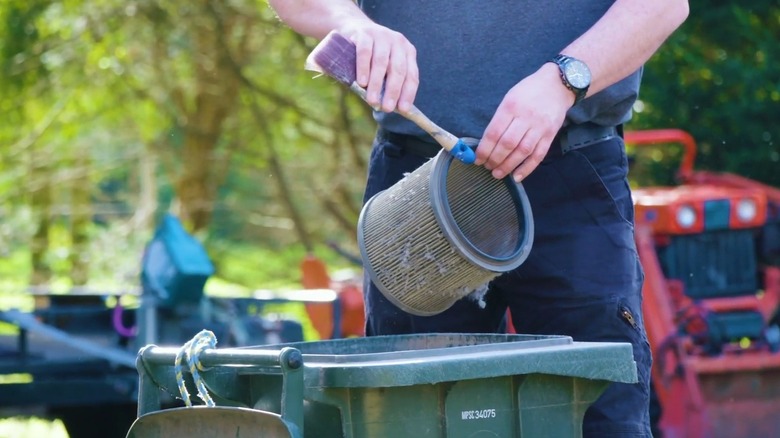 A man holding a vacuum filter over an open garbage can, cleaning out dust and debris
