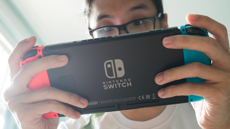 person wearing glasses holding and looking at a Nintendo Switch console in handheld mode, viewed from behind the Switch