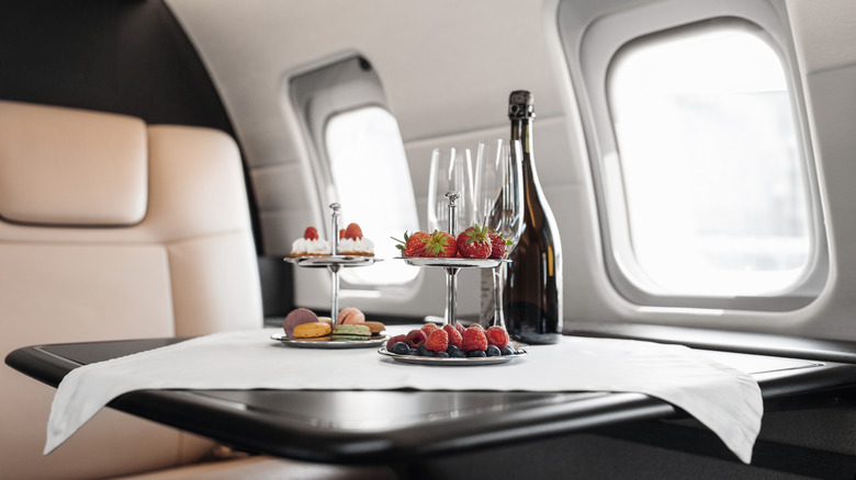 The interior of a private jet with champagne, glasses, and trays of fruit and desserts on a small table