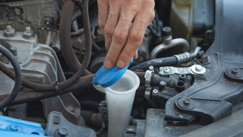 A hand opens the blue cap of a windshield washer fluid reservoir.