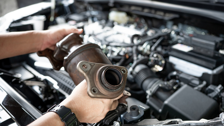 Catalytic converter held in a mechanic's hands with a car engine bay in the background