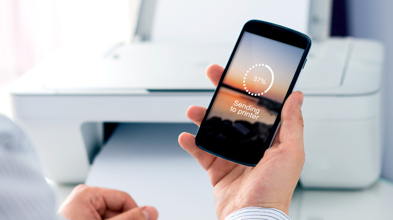 hand holding smartphone with printer in background, "Sending to Printer" displayed on the screen