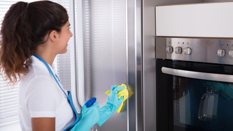A person cleaning the front of a stainless steel fridge with a cloth and spray bottle