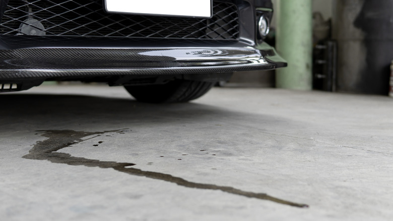 A close-up of an oil leak on a concrete floor, unidentified black car in the background