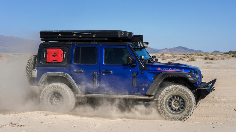 A blue Jeep drives across the desert with a bright red polypropylene fuel container attached to its side.