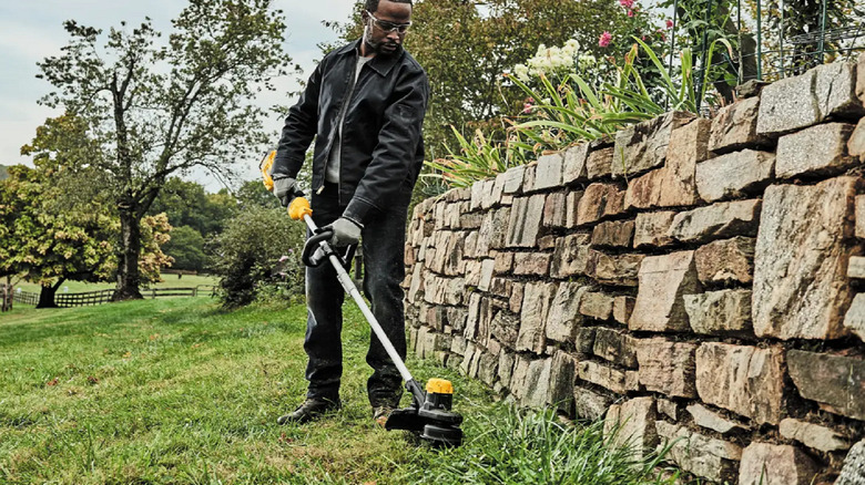 A person trimming with a DeWalt string trimmer near a cobblestone wall