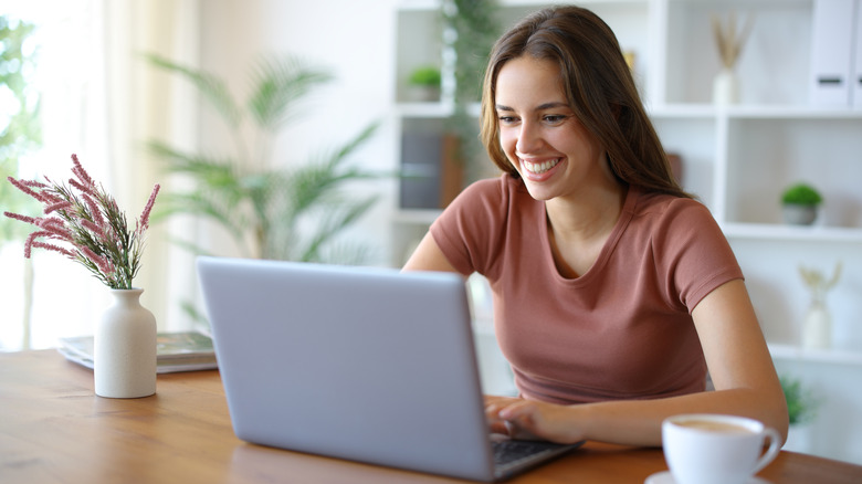 A smiling woman in a pink shirt sitting at her kitchen table with her laptop and a cup of coffee