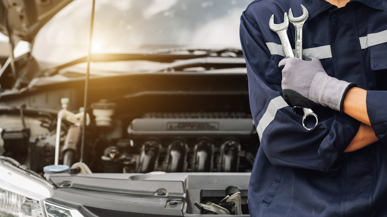 A man leaning against a car with the hood open, holding several tools