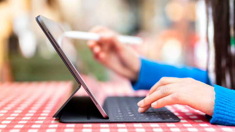 A person using an iPad with a magic keyboard and Apple Pencil on a picnic table outside