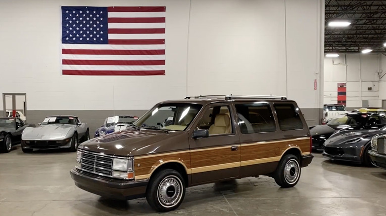 A 1990s Dodge Grand Caravan with faux woodgrain accents parked in a car dealership lot with the American flag above it.