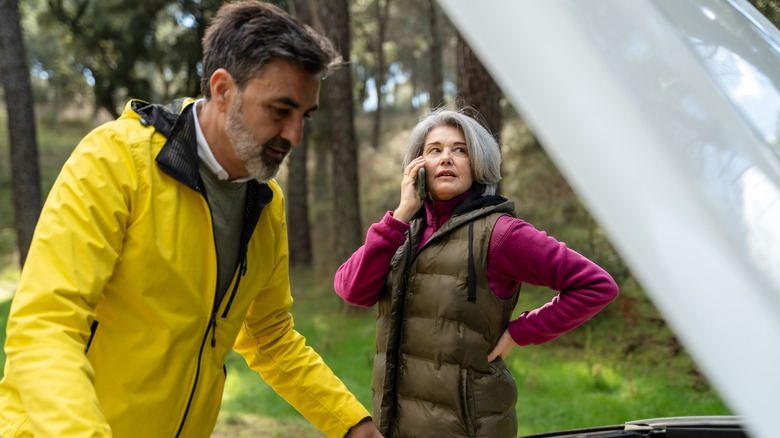 Man in yellow jacket looking under hood as woman talks on phone