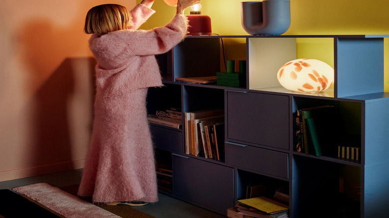 A blond woman in pink places a shade on Ikea's new orange lamp speaker, which is sitting on a shelf with another new round speaker