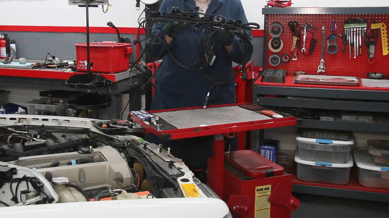 A mechanic in overalls holding up a replacement wiring harness for a cream Mercedes car in front of him with its engine bay shown.