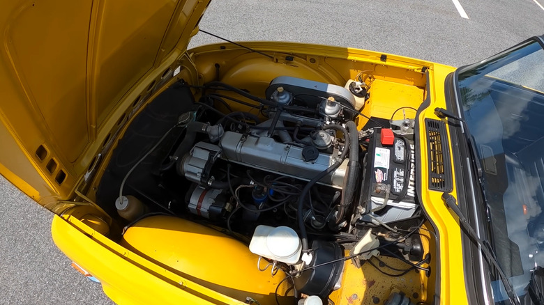 A yellow Triumph TR6 parked in a lot with its front-hinged hood open, showing its silver inline-six engine block.