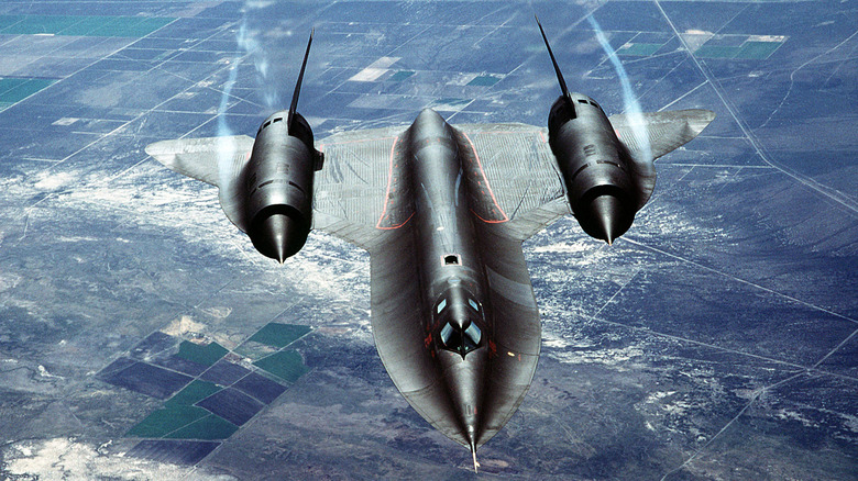 Close-up from the top of a black colored Air Force jet flying over terrain below.