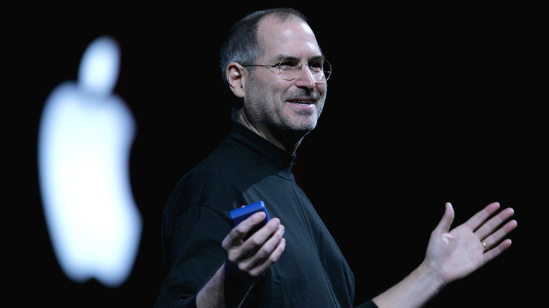Steve Jobs wearing a black turtleneck speaking in front of a lit Apple logo