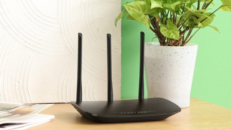 A black router with three antennae sticking up on a cluttered surface with a plant.
