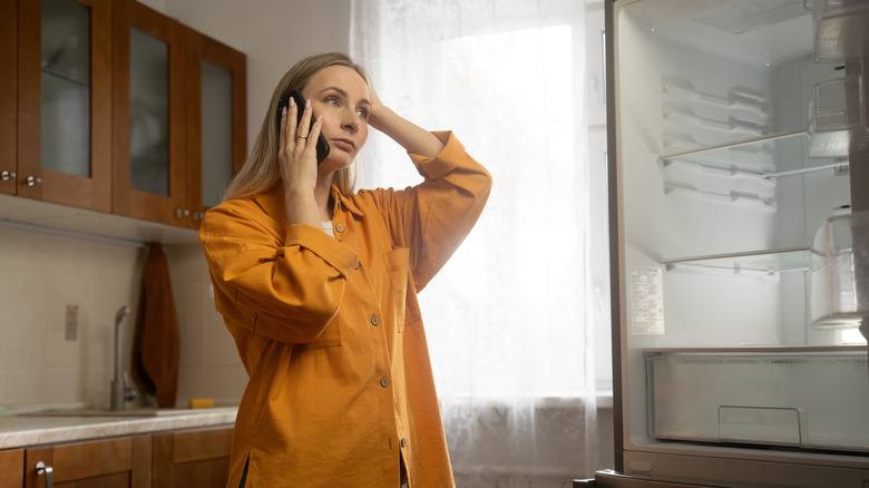 A frustrated woman in an orange shirt on the phone in front of an empty refrigerator with the door hanging open