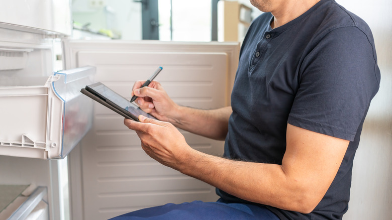A repairman kneeling in front of white refrigerator with an electronic tablet