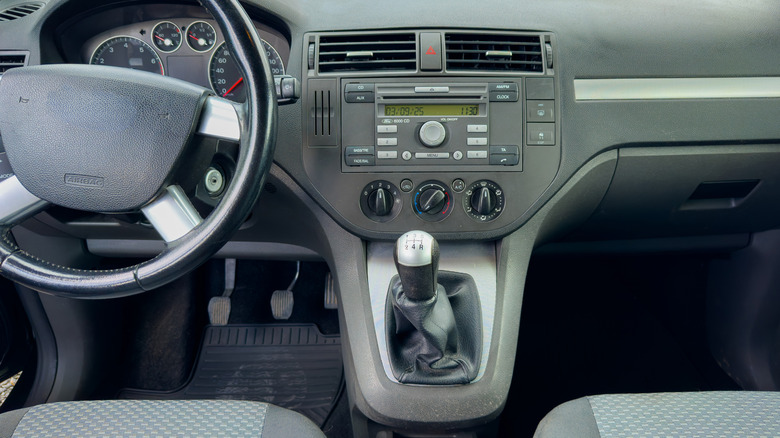 Car interior showing a steering wheel, a digital dashboard display, and a manual gear stick.