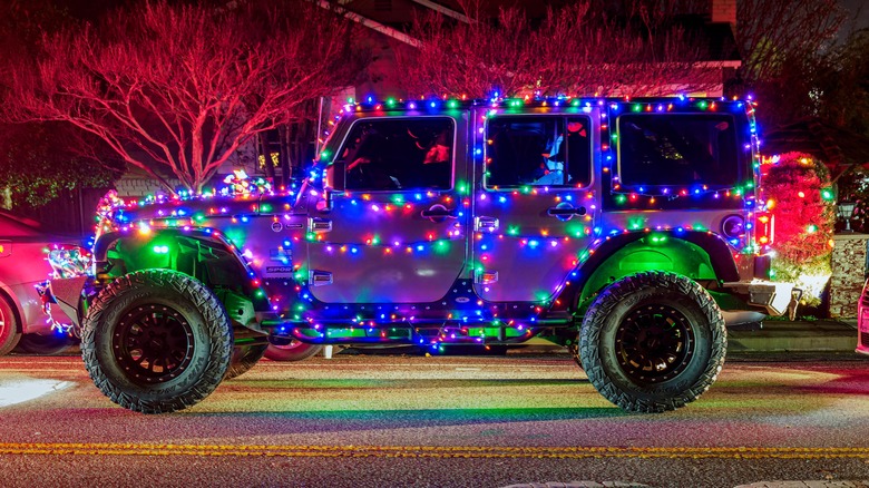 A gray Jeep Wrangler, adorned with colorful Christmas lights driving down a residential street.