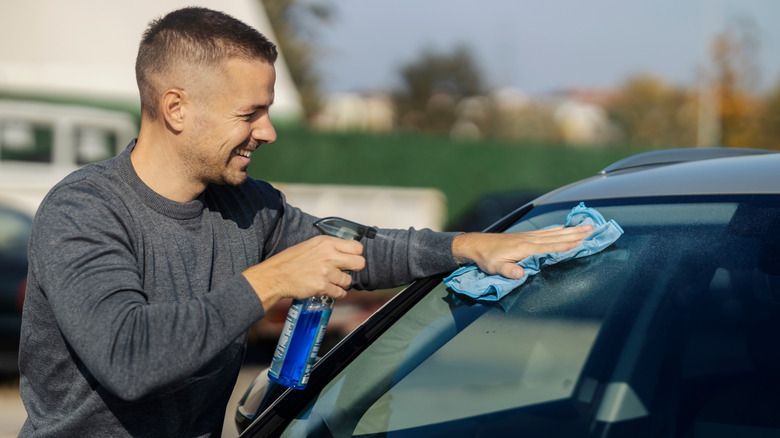 A man uses a spray cleaning product and rag on a car windshield