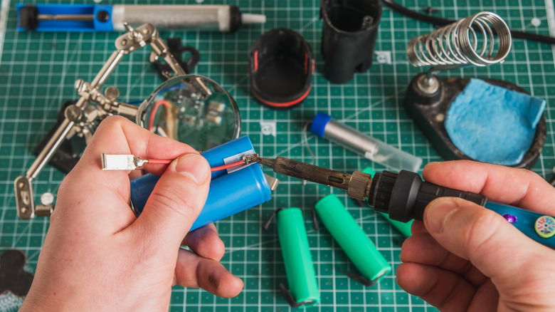 Close-up scene of repairing an old power tool battery on a soldering table with soldering iron, wires, battery cells, and green grid ma