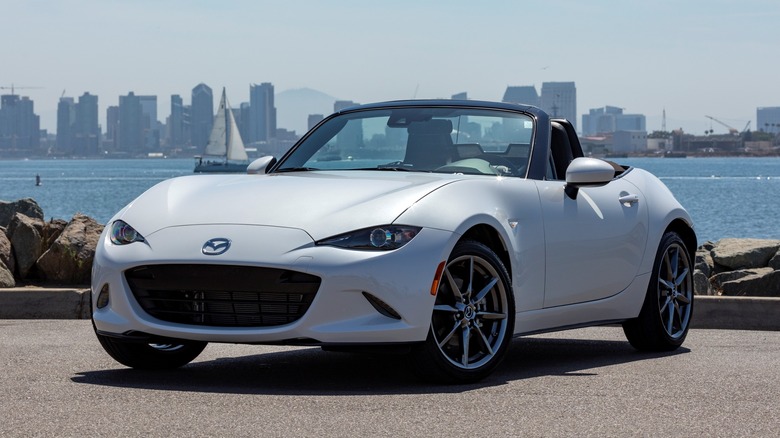 White fourth-gen Mazda Miata MX-5 parked on pavement with a body of water and city skyline in the background