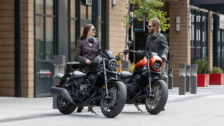 A male rider and female rider stand in front of their 2025 Honda Rebel 1100's that are parked in the city.