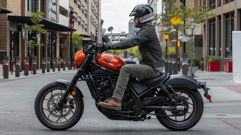 A side profile shot of a man driving an orange 2025 Honda Rebel 1100 through the city.