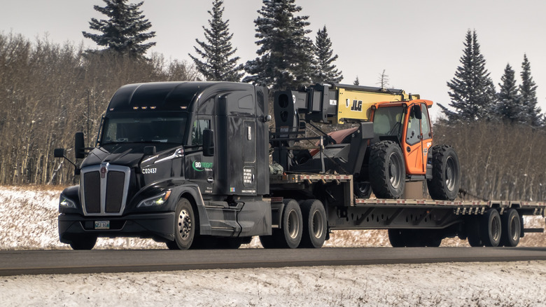 A black Kenworth T680 hauling a machine on a flatbed trailer along a highway in a snow
