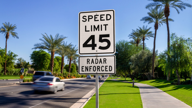 A speed limit sign on a busy four-lane road with palm trees and green grass on either side
