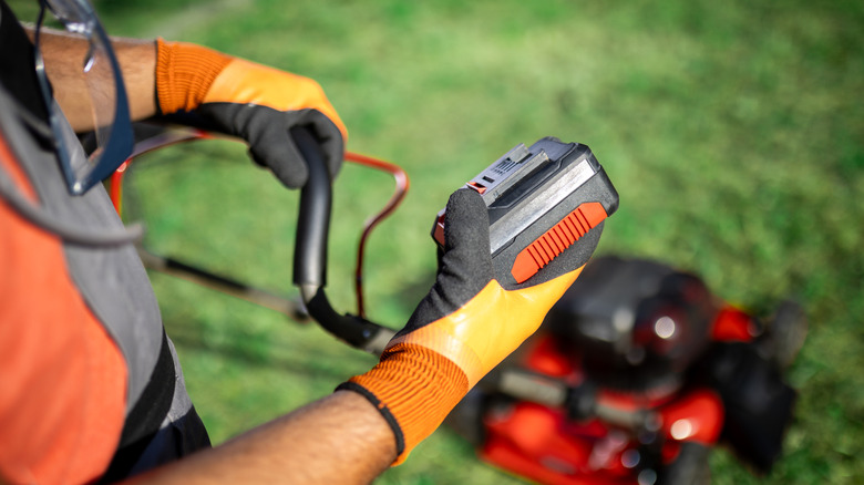 Person in gloves holds battery above lawn mower in yard