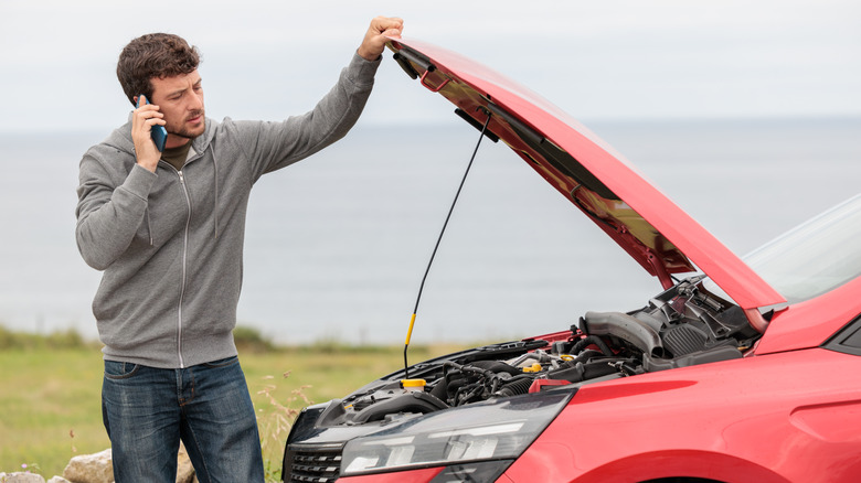 Man on the phone next to a broken down car with its hood up.