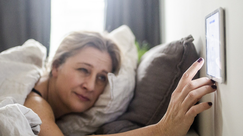A woman in bed reaching out to touch a smart thermostat on the wall