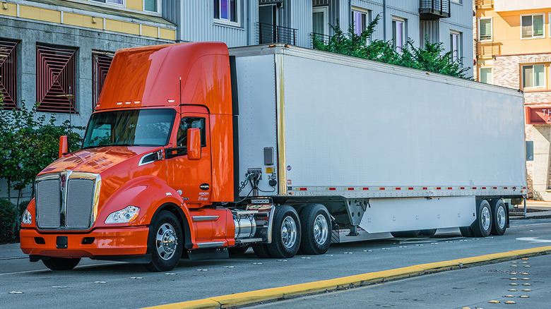 A front 3/4 shot of an orange Kenworth T680 driving through the city.