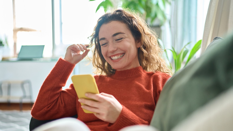 Happy relaxed young woman sitting on couch using cell phone