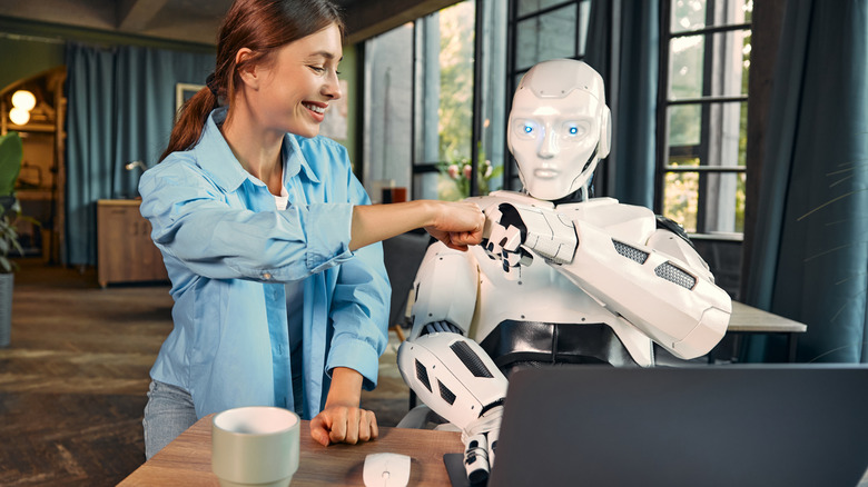 Illustration of a woman bumping fists with a humanoid robot working while sitting at a desk with a laptop.