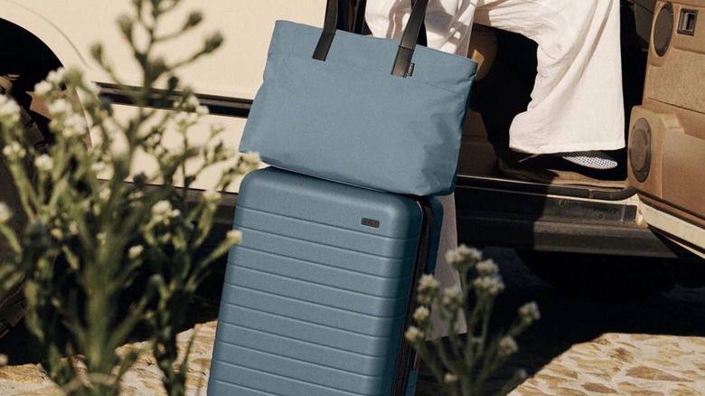 A blue Away suitcase and tote bag next to a car