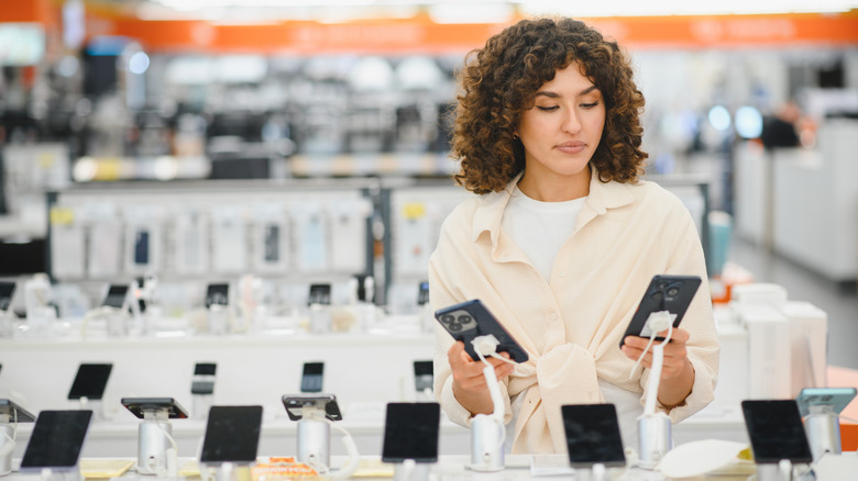 A woman with dark curly hair examines two cell phones at a display of phones in a store