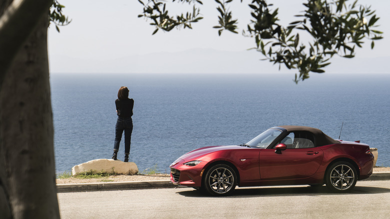 Side view of woman standing near red Mazda MX-5 Miata parked near ocean.