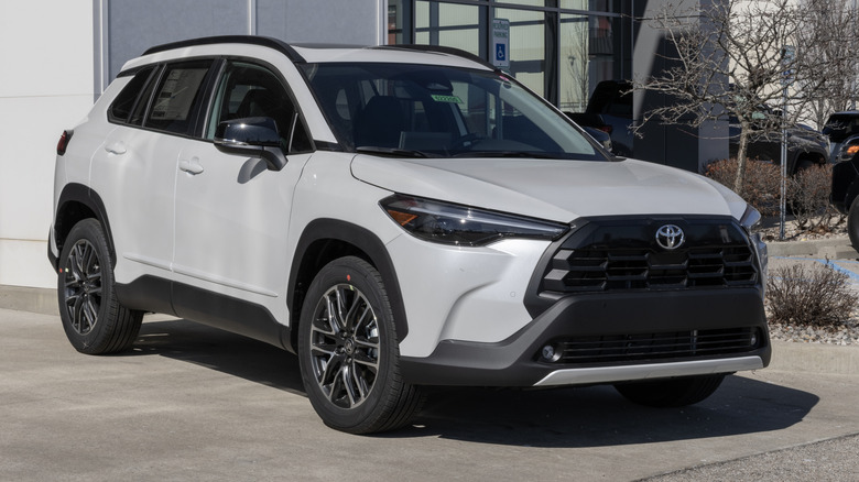 Front 3/4 view of white Toyota Corolla Cross SUV parked in front of dealership.