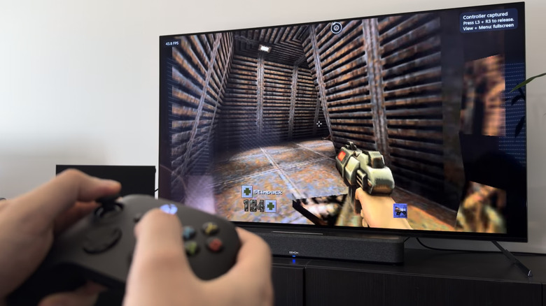 A hand holding a black Xbox controller in front of a TV running Quake